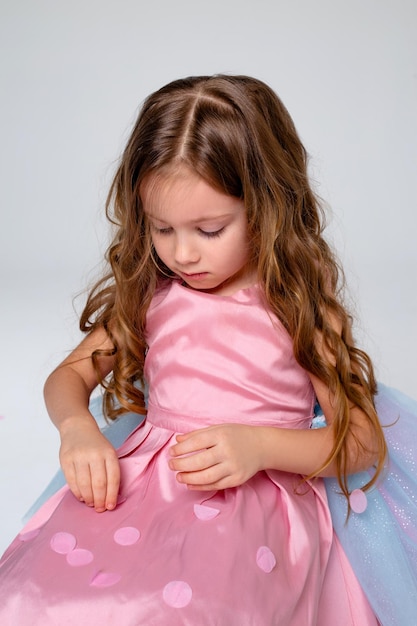 Una niña hermosa con un elegante vestido rosa posa sentada en un fondo gris y lanza confeti