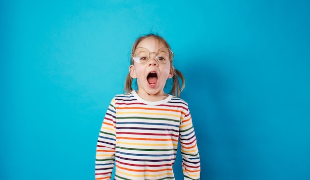 Foto una niña hermosa con gafas grandes y una camiseta a rayas se encuentra en un fondo azul aislado