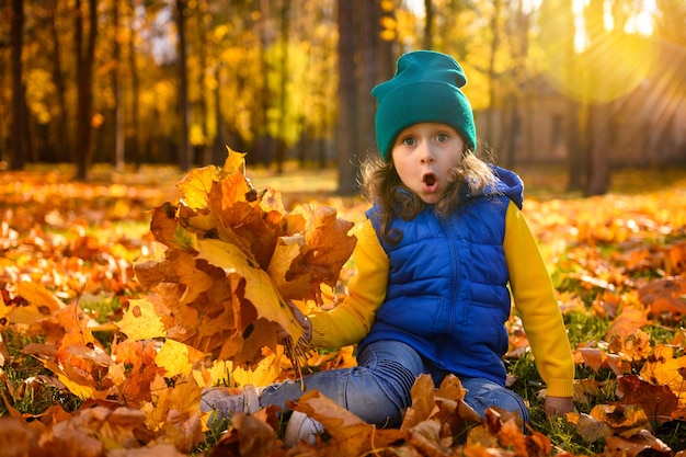 Niña hermosa y graciosa sentada en hojas caídas de arce amarillo en el bosque Niño en un paseo en el parque de otoño Niña preescolar disfrutando del clima fresco de otoño al aire libre al atardecer Estilo de vida saludable