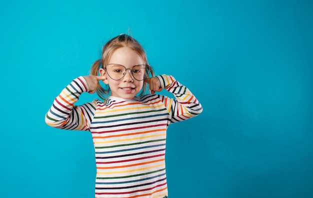 Una niña hermosa con grandes anteojos se cubre los oídos con las manos en un fondo azul aislado