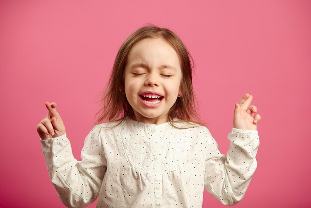 Niña hermosa con los ojos cerrados y los dedos cruzados sobre fondo rosa aislado