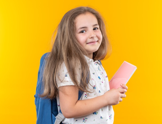 Foto niña hermosa con el pelo largo con mochila sosteniendo el cuaderno mirando al frente feliz y positivo sonriendo alegremente de pie sobre la pared naranja