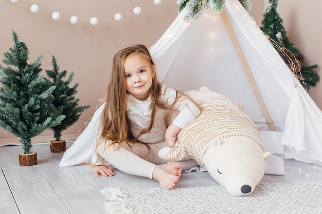 Niña hermosa en pijama blanco juega con un oso de peluche cerca del tipi. Habitación infantil con adornos y adornos para árboles de Navidad.