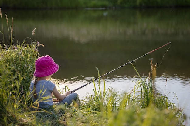 Una niña hermosa se sienta en la orilla con una caña de pescar y pesca