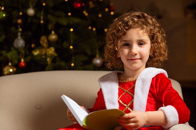 Una niña hermosa se sienta en la silla al lado de un árbol de Navidad bien decorado