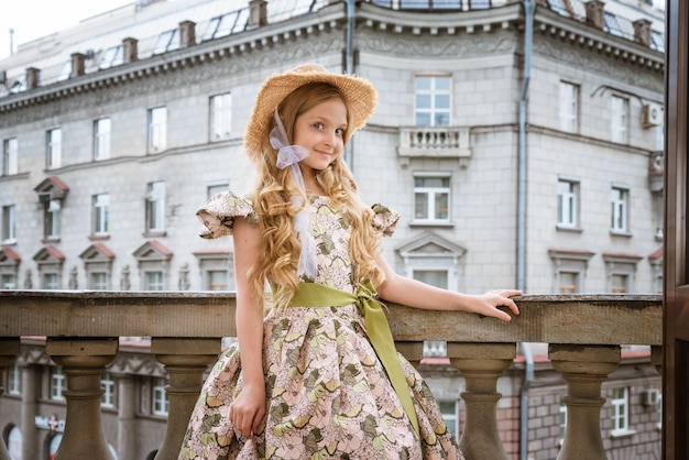 Niña hermosa en vestido y sombrero posando en el balcón de la calle