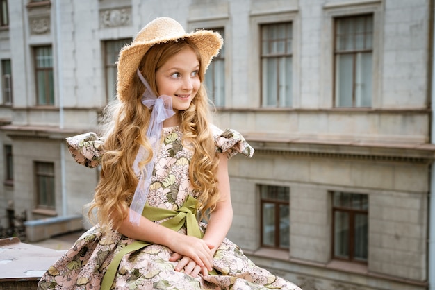 Niña hermosa en vestido y sombrero posando en el balcón de la ciudad