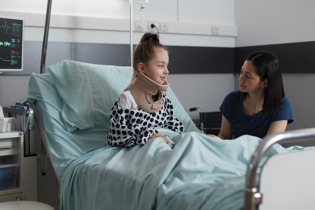 Niña hospitalizada lesionada en el cuello con collarín cervical descansando en la sala de pediatría del hospital. Niño enfermo que sufre un accidente automovilístico sentado en la cama del paciente mientras su madre la consuela.