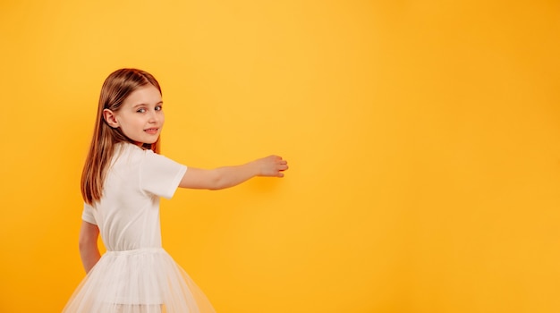 La niña imita la escritura en el escritorio y mirando a la cámara aislada sobre fondo amarillo con copyspace para su texto. Linda niña durante la sesión fotográfica de estudio