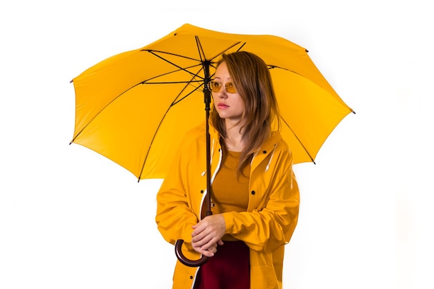 Una niña con un impermeable amarillo y gafas se encuentra debajo de un paraguas. Aislar sobre fondo blanco