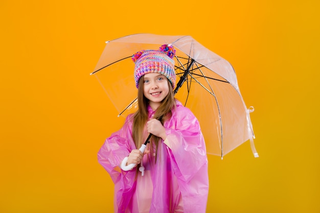 Niña en un impermeable rosa y un gorro de punto está sosteniendo un paraguas en un espacio amarillo | Premium