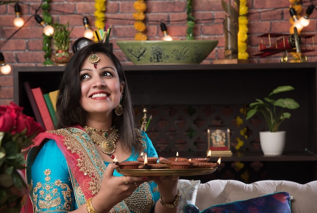 Niña india sosteniendo diwali diya en ropa tradicional, haciendo clic en la imagen selfie o hablando por teléfono inteligente.