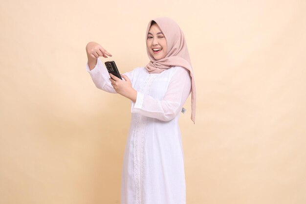 niña indonesia musulmana con hijab sonríe alegremente y hace un guiño a la cámara operando un celular pho