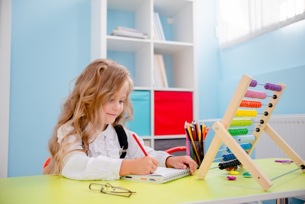 Niña inspirada en la mesa con lápices de colores. Escritorio escolar con útiles escolares, lápices, bolsos, cuaderno de apuntes y ábaco. Niña rubia con gafas de nuevo al concepto de escuela.