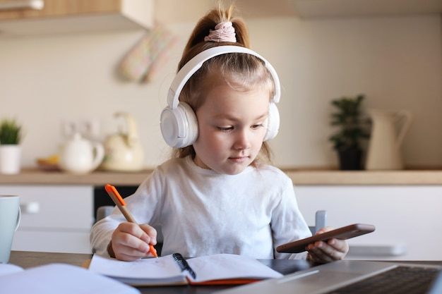 La niña inteligente con auriculares hace la clase de tarea en línea en el teléfono inteligente, el niño pequeño con auriculares estudia en Internet, tiene una conferencia web o una lección durante la cuarentena.