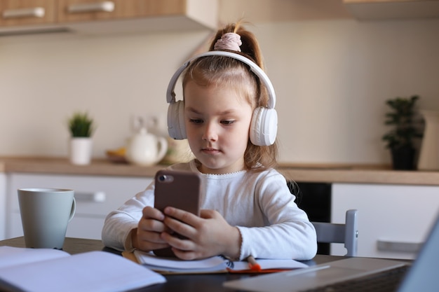 La niña inteligente con auriculares hace la clase de tarea en línea en el teléfono inteligente, el niño pequeño con auriculares estudia en Internet, tiene una conferencia web o una lección durante la cuarentena.
