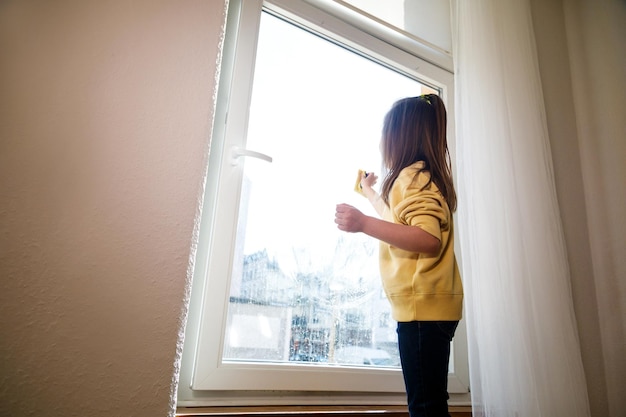 Niña interior con guantes amarillos lavando ventanas en primavera