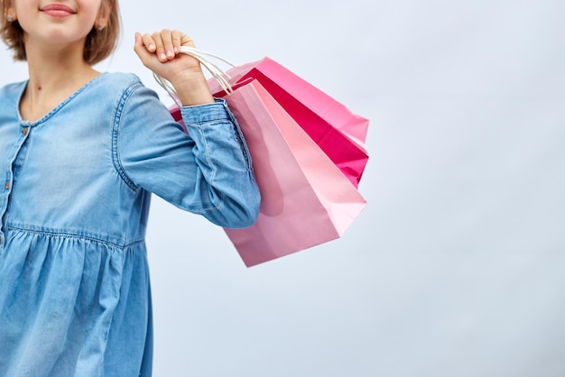 Niña irreconocible en vestido de mezclilla casual con bolsas de compras rosa en la mano