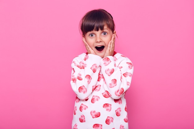 Niña en jersey blanco con corazones, tiene noticias de última hora, cubriendo las mejillas con las palmas, mirando a la cámara con expresión de sorpresa, aislada sobre una pared rosa.