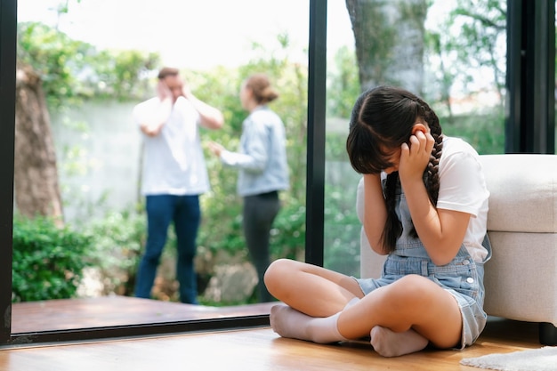 Niña joven estresada e infeliz acurrucada en una esquina cubriéndose las orejas bloqueando el sonido de sus padres discutiendo en el fondo La violencia doméstica en el hogar y la infancia traumática se desarrollan en depresión Sincronías