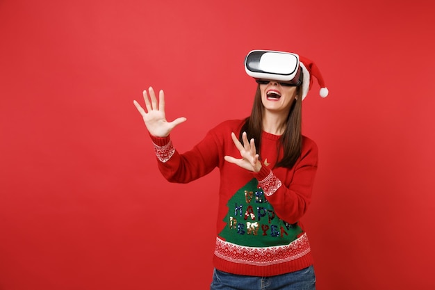 La niña joven de Santa mirando en el auricular toca algo como presionar el botón, apuntando a la pantalla virtual flotante aislada sobre fondo rojo. Feliz año nuevo 2019 celebración concepto de fiesta navideña.