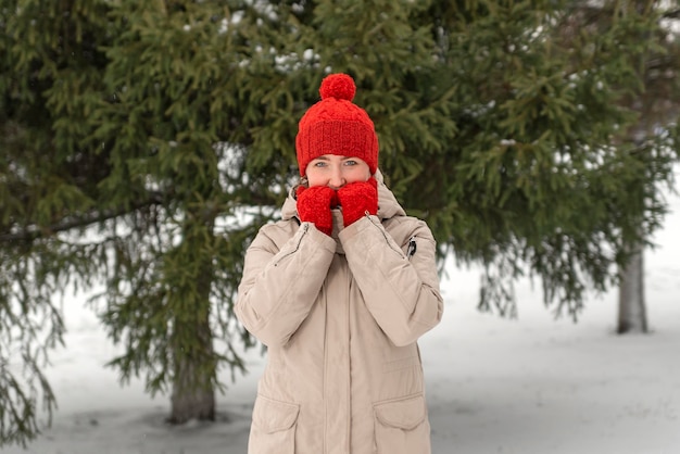 Foto niña joven con sombrero de lana roja y guantes calienta las manos de pie cerca del pino verde en el parque de invierno o el bosque paseo de invierno