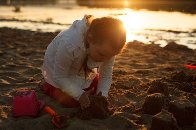 Una niña juega con arena en una playa de arena en la orilla del río durante la puesta de sol.