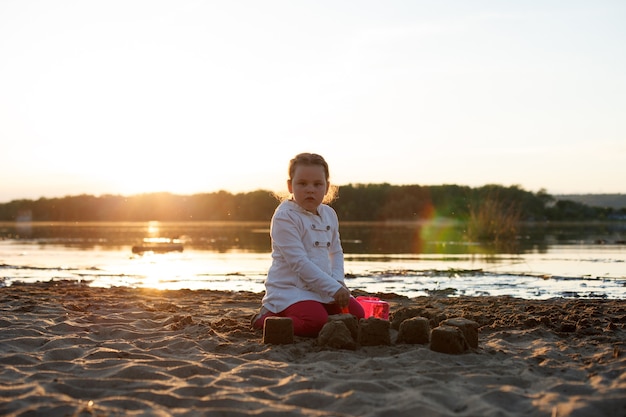 Una niña juega con arena en una playa de arena en la orilla del río durante la puesta de sol.