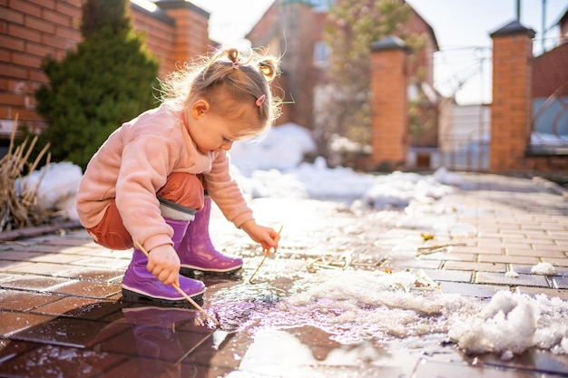 Niña juega en un charco con palo de madera en primavera a la luz del sol