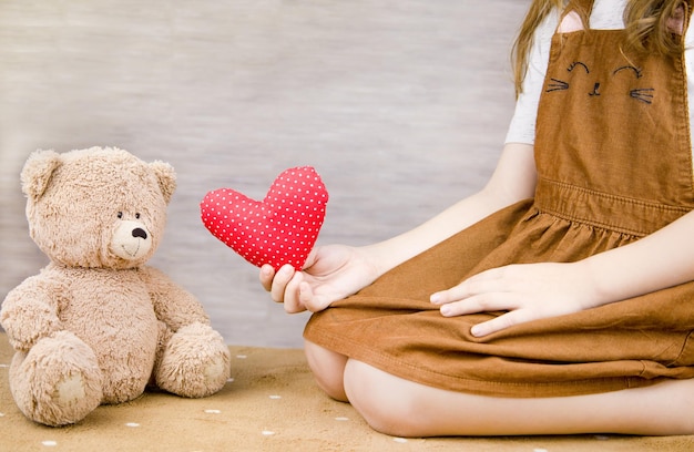 Una niña juega con un osito de peluche. El niño tiene un corazón rojo en las manos.