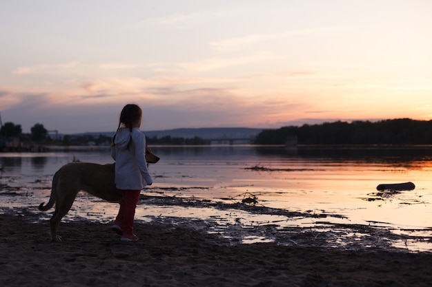 Una niña juega con un perro en una playa de arena junto al río al atardecer.