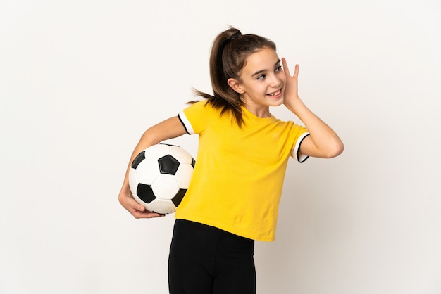 Niña de jugador de fútbol aislado en la pared blanca escuchando algo poniendo la mano en la oreja