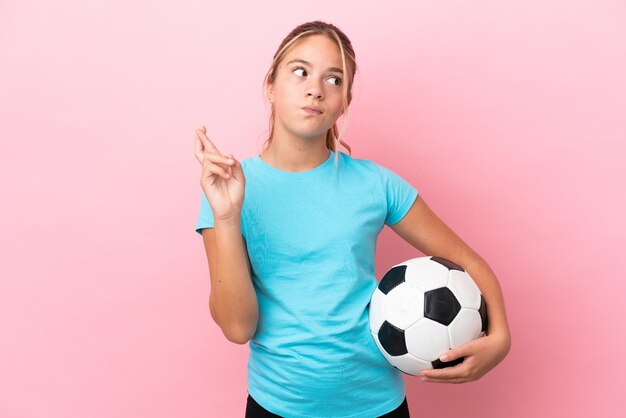 Niña de jugador de fútbol aislado sobre fondo rosa con los dedos cruzando y deseando lo mejor