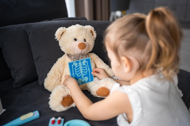 Niña jugando al doctor con juguetes y oso de peluche en el sofá en la sala de estar en casa