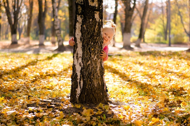 Niña jugando al escondite se asoma desde detrás de un árbol de fondo de otoño