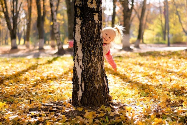 Niña jugando al escondite se asoma desde detrás de un árbol de fondo de otoño