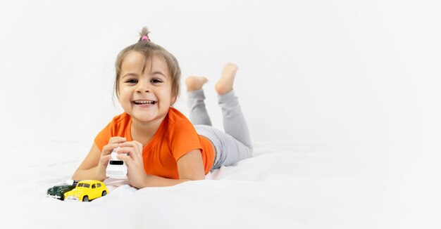 Niña jugando con coche de juguete blanco. Sentado en la sábana blanca. Sonriente. Niños en edad preescolar.