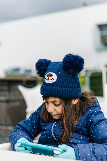 Niña jugando consola portátil, con ropa de invierno azul y gorro de lana