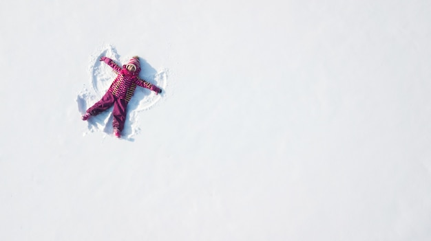 Niña jugando y haciendo un ángel de nieve en la nieve. Vista superior plana desde arriba