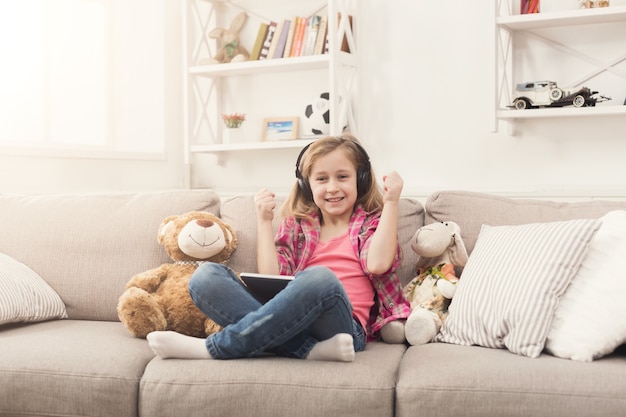 Niña jugando juegos en línea en tableta digital y escuchando música en auriculares. Niña sentada en el sofá con sus juguetes. Concepto de educación en línea y redes sociales