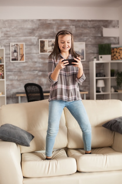 Niña jugando juegos en teléfonos inteligentes saltando en el sofá de la sala de estar.