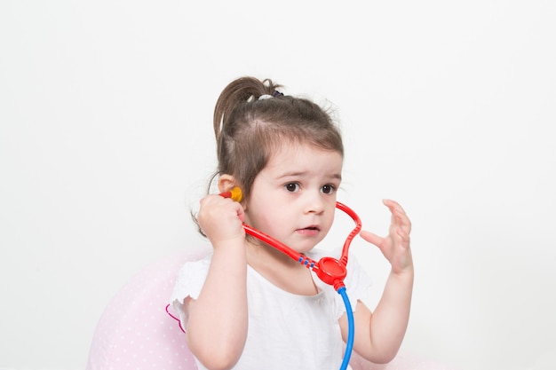 Niña jugando a un médico con estetoscopio de juguete sobre fondo blanco.