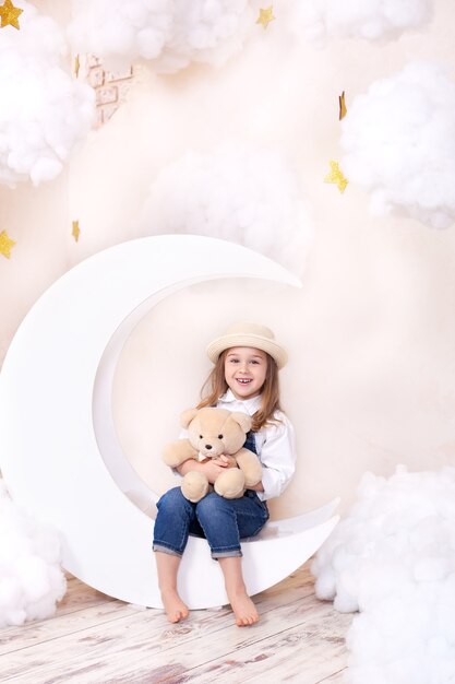 Niña jugando con osito de peluche sentado en una luna en la habitación con nubes