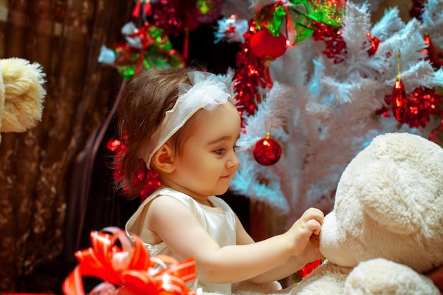 Niña jugando con su osito de peluche bajo el árbol de Navidad blanco