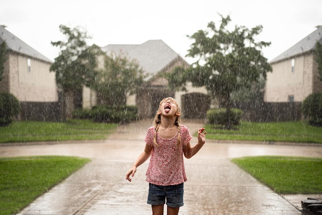 Niña juguetona con la boca abierta de pie en el camino de entrada durante la lluvia