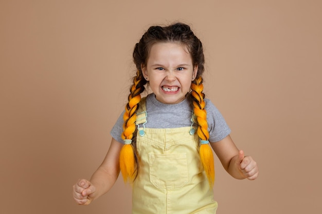 Niña juguetona y complaciente con coletas amarillas de kanekalon asustando a la gente con dientes descubiertos con dientes faltantes usando mono amarillo y camiseta gris sobre fondo beige