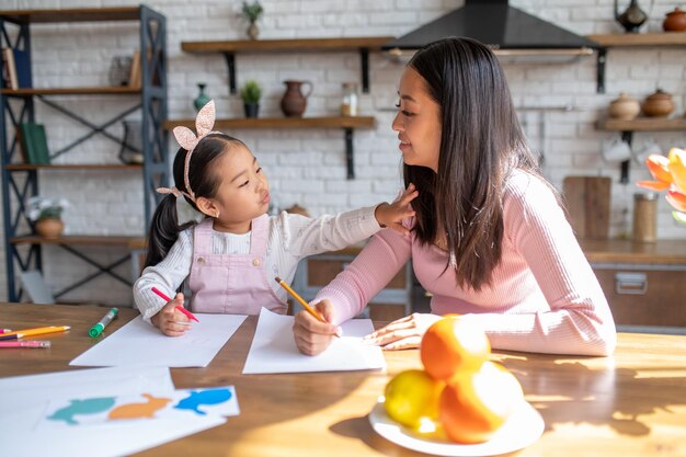 Niña juguetona con un marcador de color y su madre con un lápiz sentados juntos en la mesa de la cocina