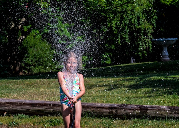 Niña juguetona en traje de baño rociando agua con manguera de jardín en el césped