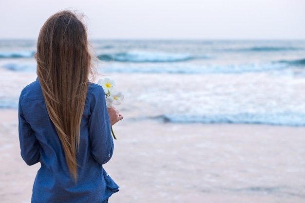 Una niña junto al mar al atardecer, sosteniendo una flor de plumeria en su mano