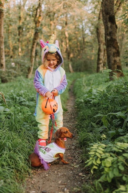 Niña en un kigurumi y un perro salchicha enano en disfraces de Halloween están caminando en el bosque